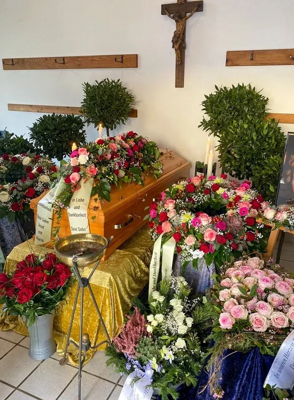 Heller Sarg mit Blumengesteck in der Trauerhalle, an der Wand ein Kruzifix, flankiert von Lorbeerbäumchen und Kerzen.