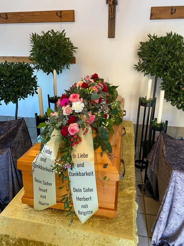 Heller Holzsarg mit einem Gesteck aus rosa und roten Rosen, flankiert von Lorbeerbäumchen unter einem Kruzifix.
