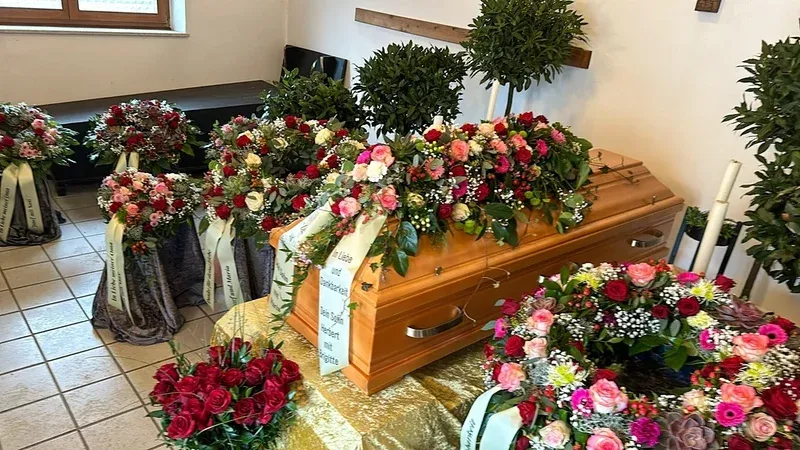 Heller Sarg in der Trauerhalle, umgeben von zahlreichen Gestecken aus roten und rosa Rosen.
