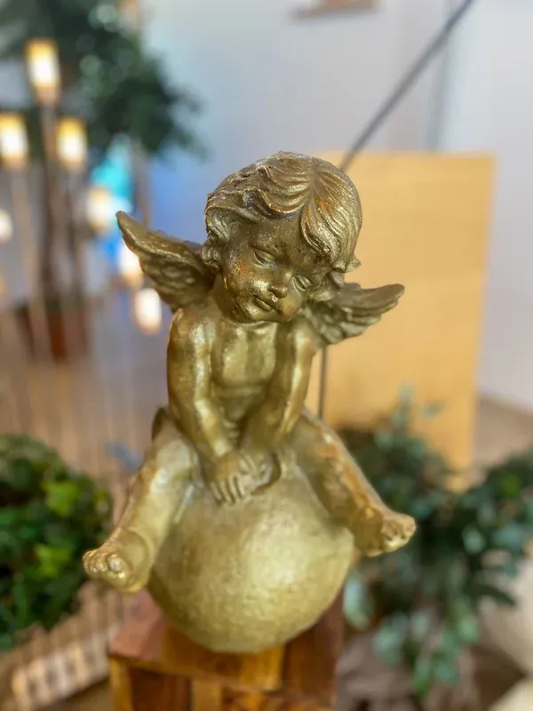 Nachdenkliche goldene Engelsfigur auf einer Kugel, im Hintergrund warme Kerzenlichter.