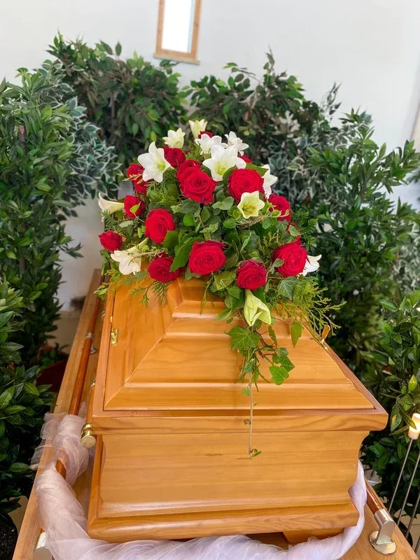 Heller Holzsarg mit einem Gesteck aus roten Rosen und weißen Lilien, umgeben von grünen Pflanzen.