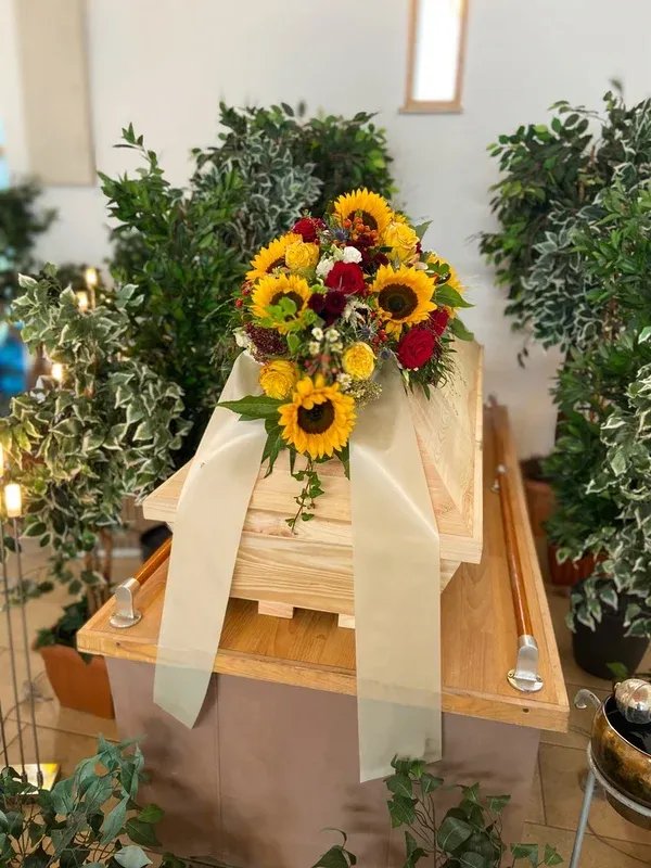 Heller Holzsarg mit einem sommerlichen Gesteck aus Sonnenblumen und roten Rosen.