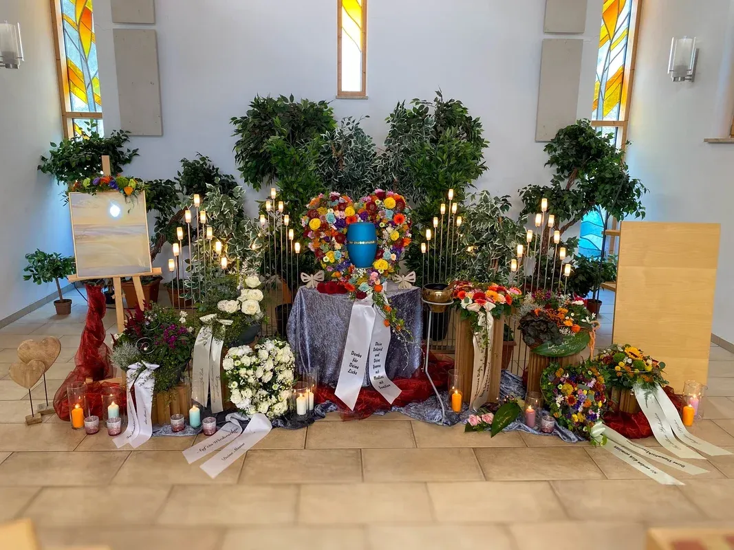 Festlich gestalteter Abschiedsraum: eine blaue Urne in einem großen Blütenherz, umgeben von Kerzen und Gestecken vor Buntglasfenstern.