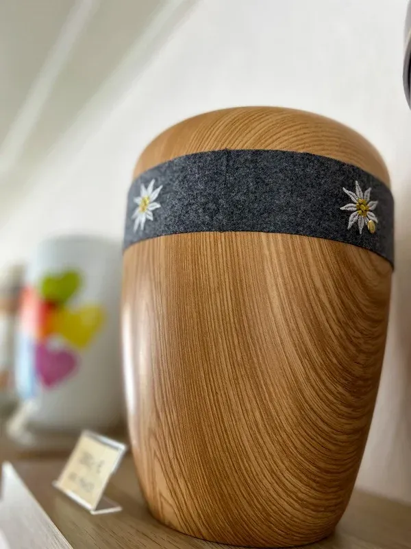 Urne aus hellem Holz mit einem Filzband und Edelweiß-Stickerei in der Ausstellung.