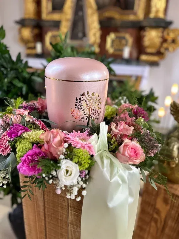 Rosafarbene Urne mit goldenem Baummotiv, umgeben von einem Gesteck aus rosa Rosen und Gerbera vor einem goldenen Altar.