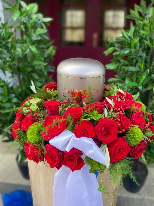 Silberne Urne, dicht umkränzt von roten Rosen, in Nahaufnahme vor einem roten Portal.