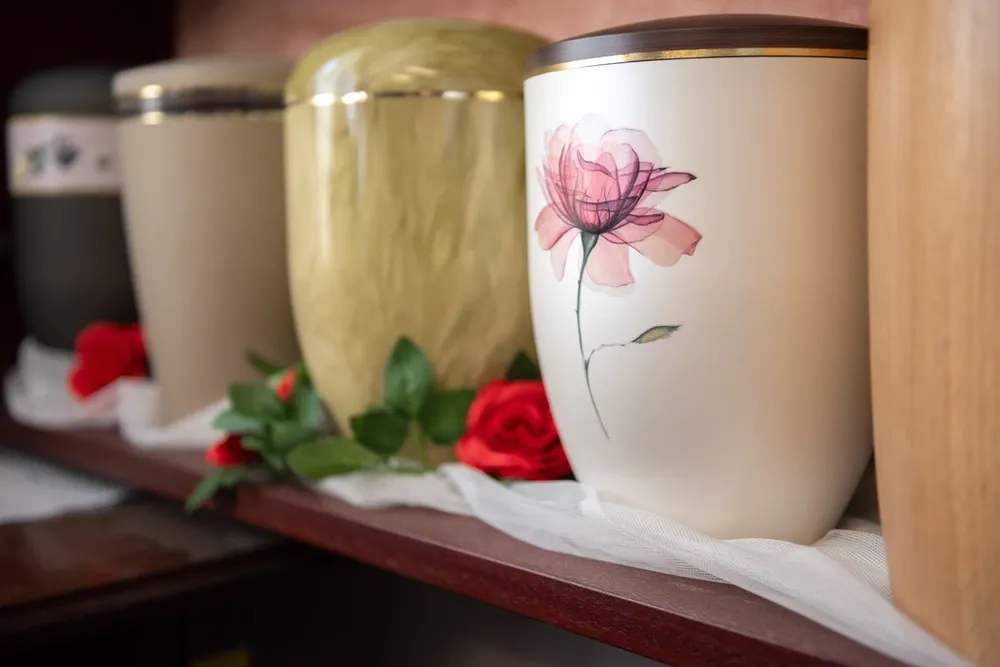 Weiße Urne mit zartem Blütenmotiv im Regal, daneben weitere Urnen und rote Rosen.