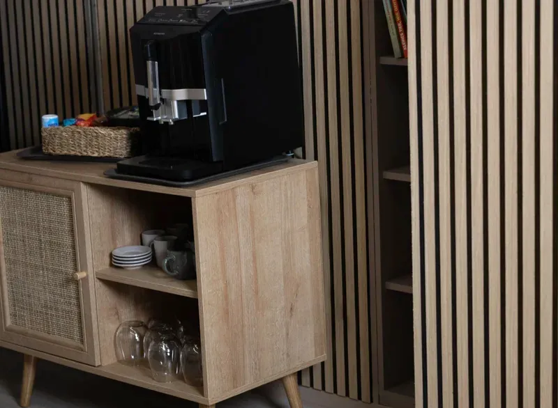 Kaffeevollautomat mit Tassen und Gläsern auf einem Sideboard vor einer Akustik-Holzwand im Wartebereich.